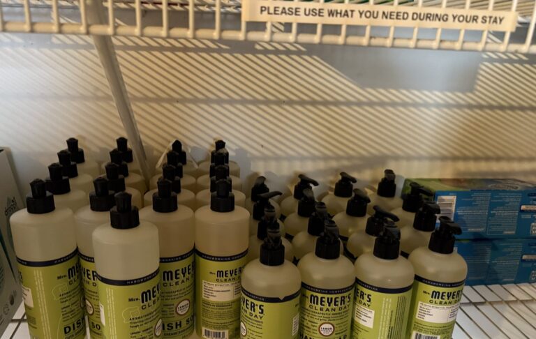 Shelf stocked with rows of Mrs. Meyer's hand soap and dish soap for guests, beneath a sign reading Please use what you need during your stay