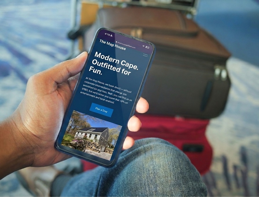 A traveler seated by luggage in an airport holds a phone showing the Map House vacation rental homepage, headline Modern Cape Outfitted for Fun with a Plan a Stay button