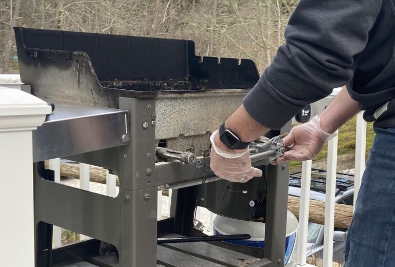 Gloved hands repairing the disassembled burner assembly of an outdoor grill taken apart on its cart