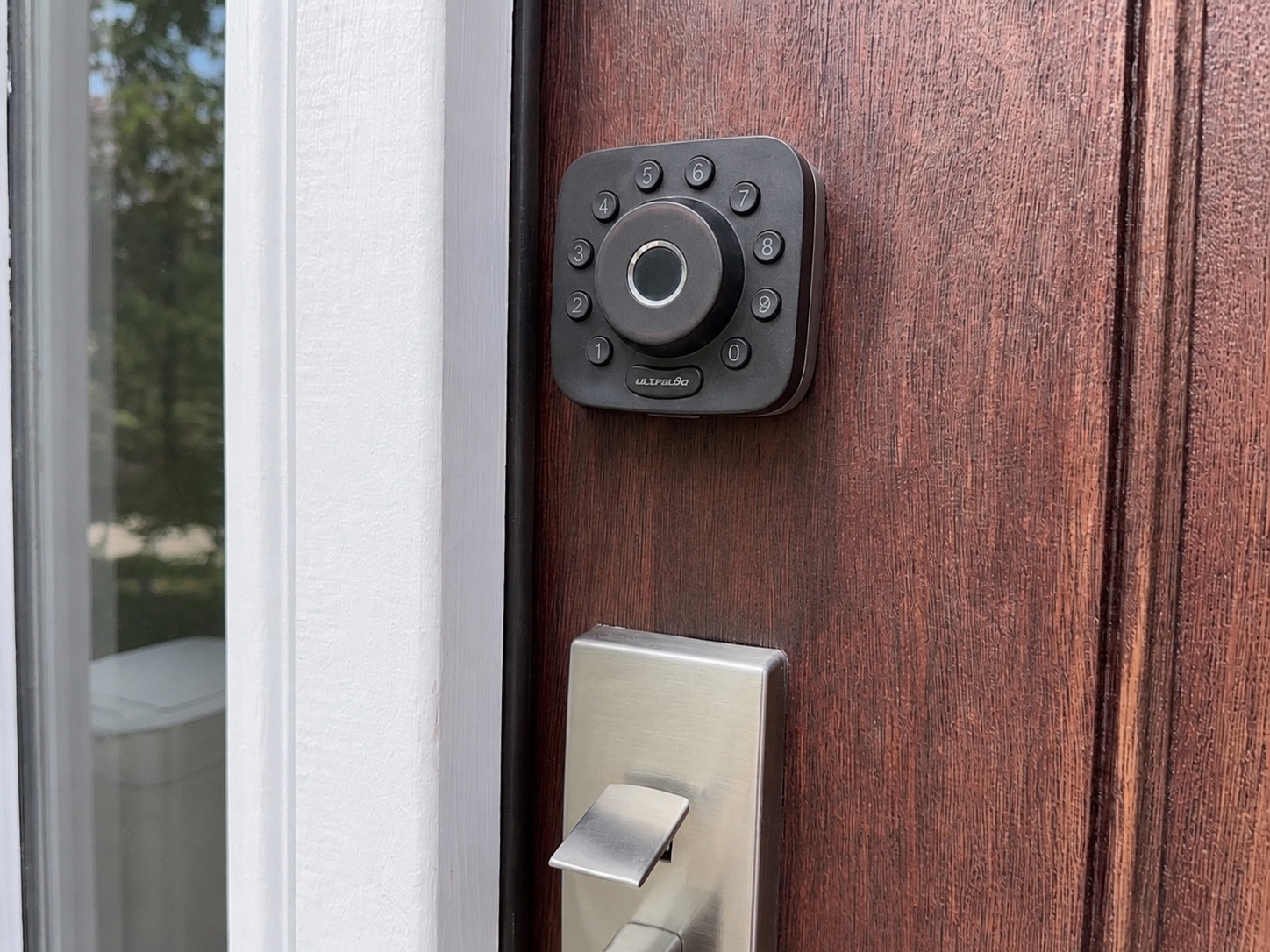 An Ultraloq keypad and fingerprint smart lock mounted on a wood front door above a brushed-nickel lever handle